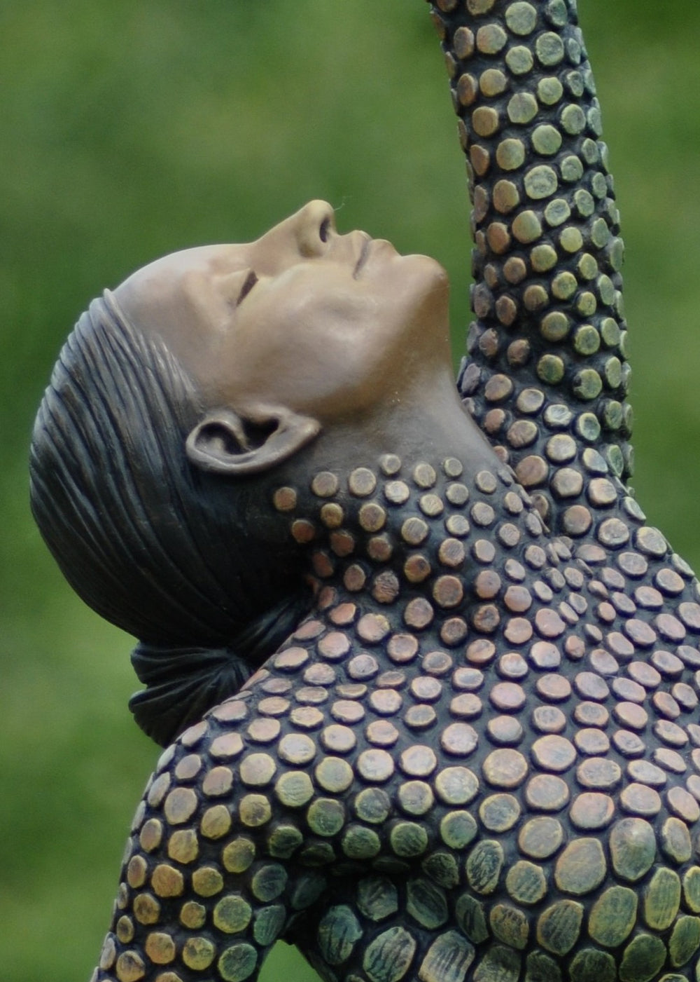 Life-size bronze sculpture of a woman balancing on one foot, her body covered in hand-painted mosaic-like textured scales that shift from earthy tones to green and blue patinas. She reaches upward with eyes closed, captured in a graceful, expressive pose against an outdoor garden backdrop.