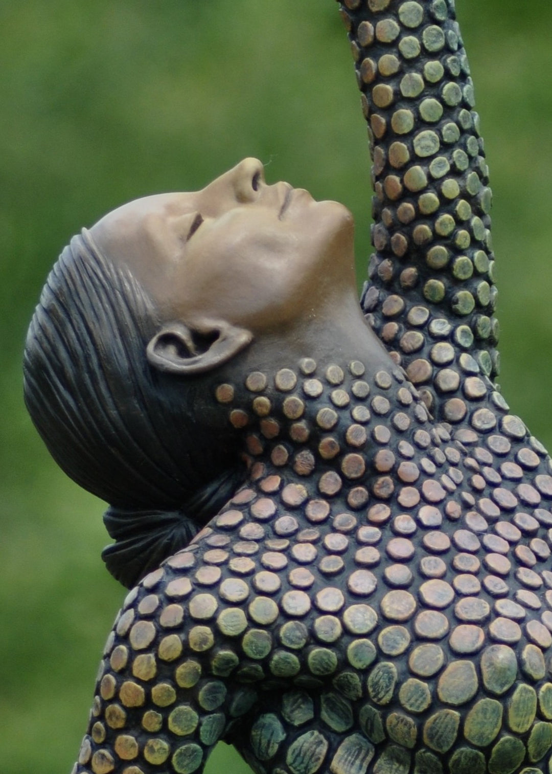 Life-size bronze sculpture of a woman balancing on one foot, her body covered in hand-painted mosaic-like textured scales that shift from earthy tones to green and blue patinas. She reaches upward with eyes closed, captured in a graceful, expressive pose against an outdoor garden backdrop.