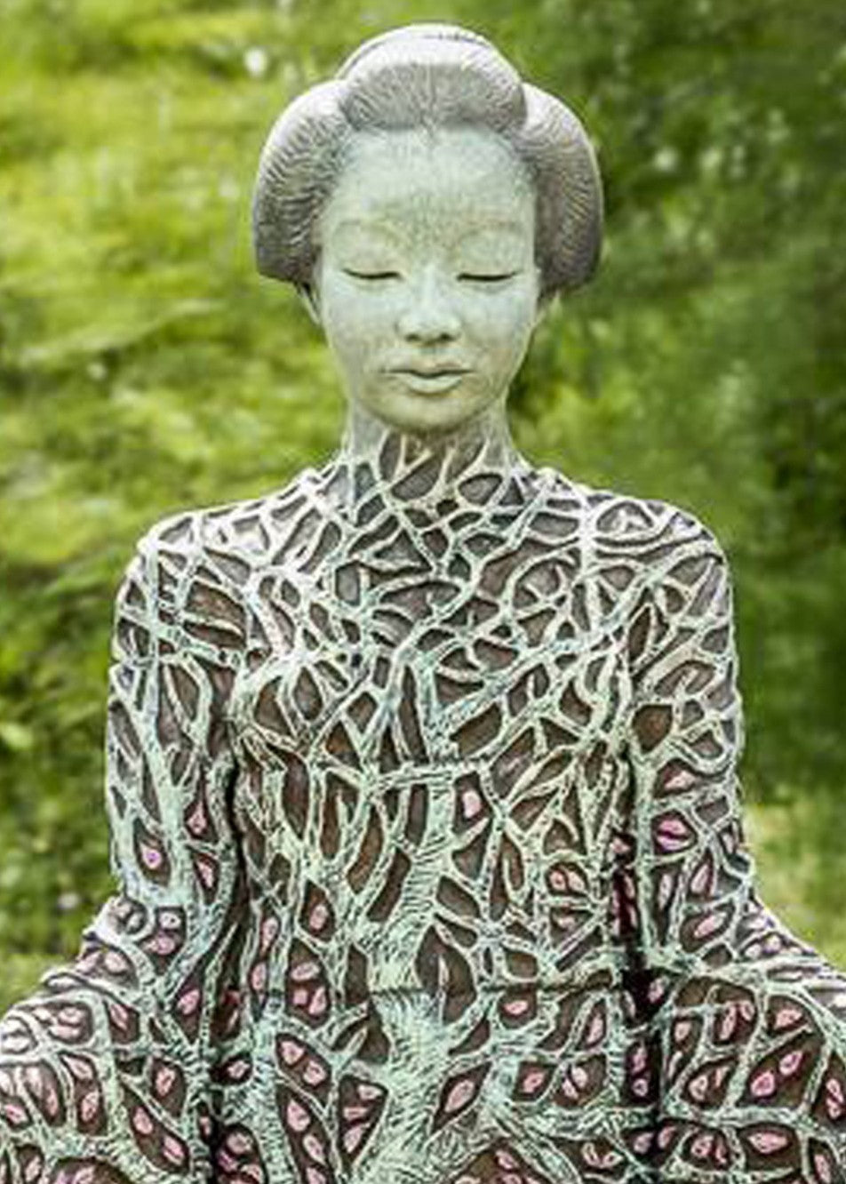 Bronze sculpture of a serene female figure standing with softly extended arms, her body intricately patterned as a blossoming tree. Pale green branches and trunk flow through her torso, while hundreds of delicate pink leaves form her robe and cascade toward the ground. The sculpture stands outdoors, surrounded by soft, blurred greenery that enhances its tranquil, meditative presence.