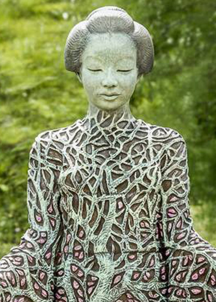 Bronze sculpture of a serene female figure standing with softly extended arms, her body intricately patterned as a blossoming tree. Pale green branches and trunk flow through her torso, while hundreds of delicate pink leaves form her robe and cascade toward the ground. The sculpture stands outdoors, surrounded by soft, blurred greenery that enhances its tranquil, meditative presence.