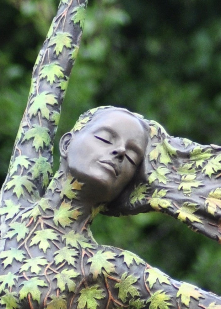 Bronze sculpture of a female figure balancing gracefully on one foot, her body and flowing dress covered in hand-painted maple leaves that shift from fresh greens to warm yellows, oranges and deep autumnal reds. She leans into the wind with one arm raised, hair swept back, standing outdoors against dense green foliage.