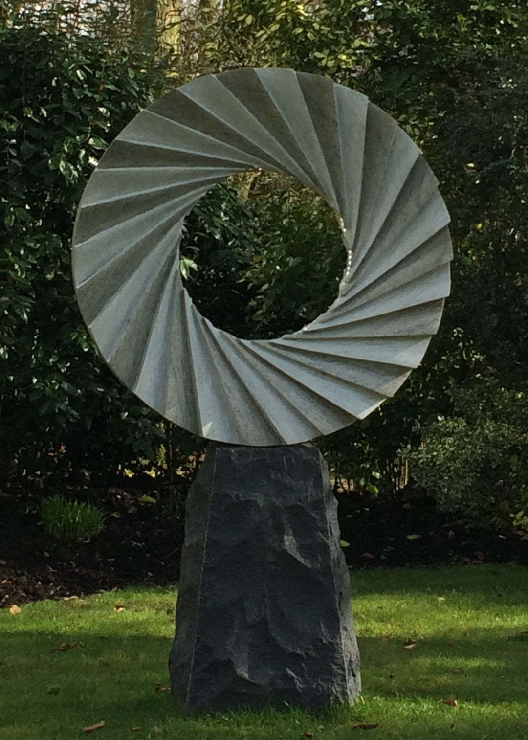 ‘Catherine Wheel VIII’ by Nicolas Moreton MRSS, a unique contemporary abstract stone sculpture carved from Ancaster Weatherbed Limestone, featuring a large circular form with a central opening surrounded by precisely carved, twisting radial ridges that create a spiralling pattern. The pale grey and cream limestone contrasts with the rough, dark charcoal stone pedestal. Integrated LED lighting illuminates the inner edges of the spiral with bright white light, creating alternating bands of light and shadow. P