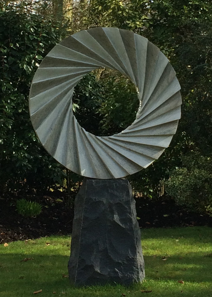 ‘Catherine Wheel VIII’ by Nicolas Moreton MRSS, a unique contemporary abstract stone sculpture carved from Ancaster Weatherbed Limestone, featuring a large circular form with a central opening surrounded by precisely carved, twisting radial ridges that create a spiralling pattern. The pale grey and cream limestone contrasts with the rough, dark charcoal stone pedestal. Integrated LED lighting illuminates the inner edges of the spiral with bright white light, creating alternating bands of light and shadow. P
