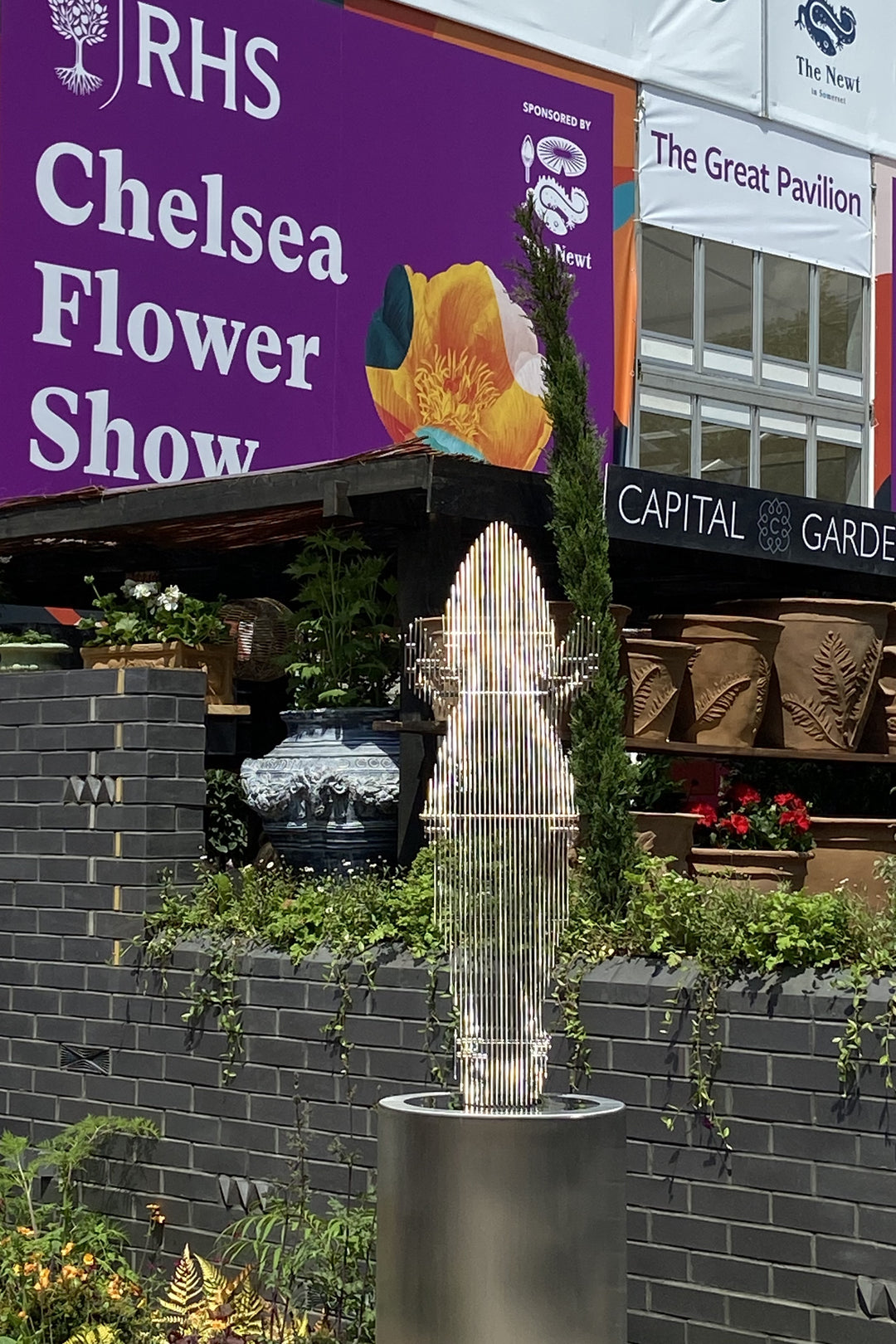 A sculptural stainless steel horse head by Tom Hiscox, featuring flowing contours and stylised form, displayed outdoors as a freestanding installation amid lush garden planting at the RHS Chelsea Flower Show, with rear views revealing the vertical steel slices and internal structure, culminating in a striking illuminated presentation set against the event’s floral and architectural backdrop.