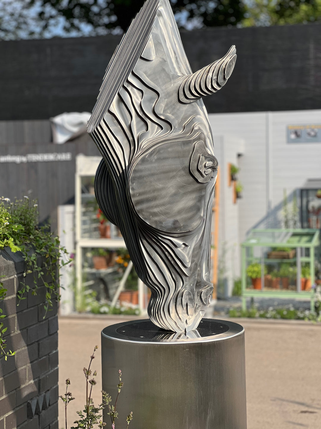 A sculptural stainless steel horse head by Tom Hiscox, featuring flowing contours and stylised form, displayed outdoors as a freestanding installation amid lush garden planting at the RHS Chelsea Flower Show, with rear views revealing the vertical steel slices and internal structure, culminating in a striking illuminated presentation set against the event’s floral and architectural backdrop.