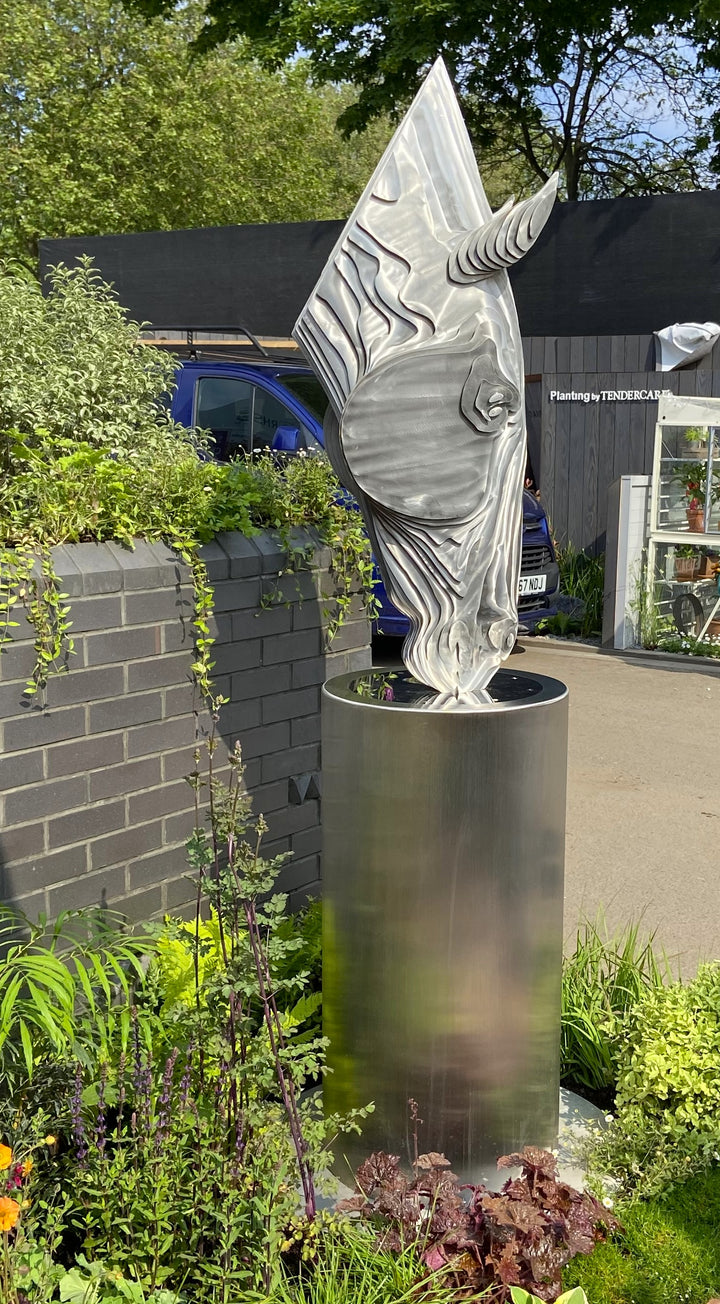 A sculptural stainless steel horse head by Tom Hiscox, featuring flowing contours and stylised form, displayed outdoors as a freestanding installation amid lush garden planting at the RHS Chelsea Flower Show, with rear views revealing the vertical steel slices and internal structure, culminating in a striking illuminated presentation set against the event’s floral and architectural backdrop.