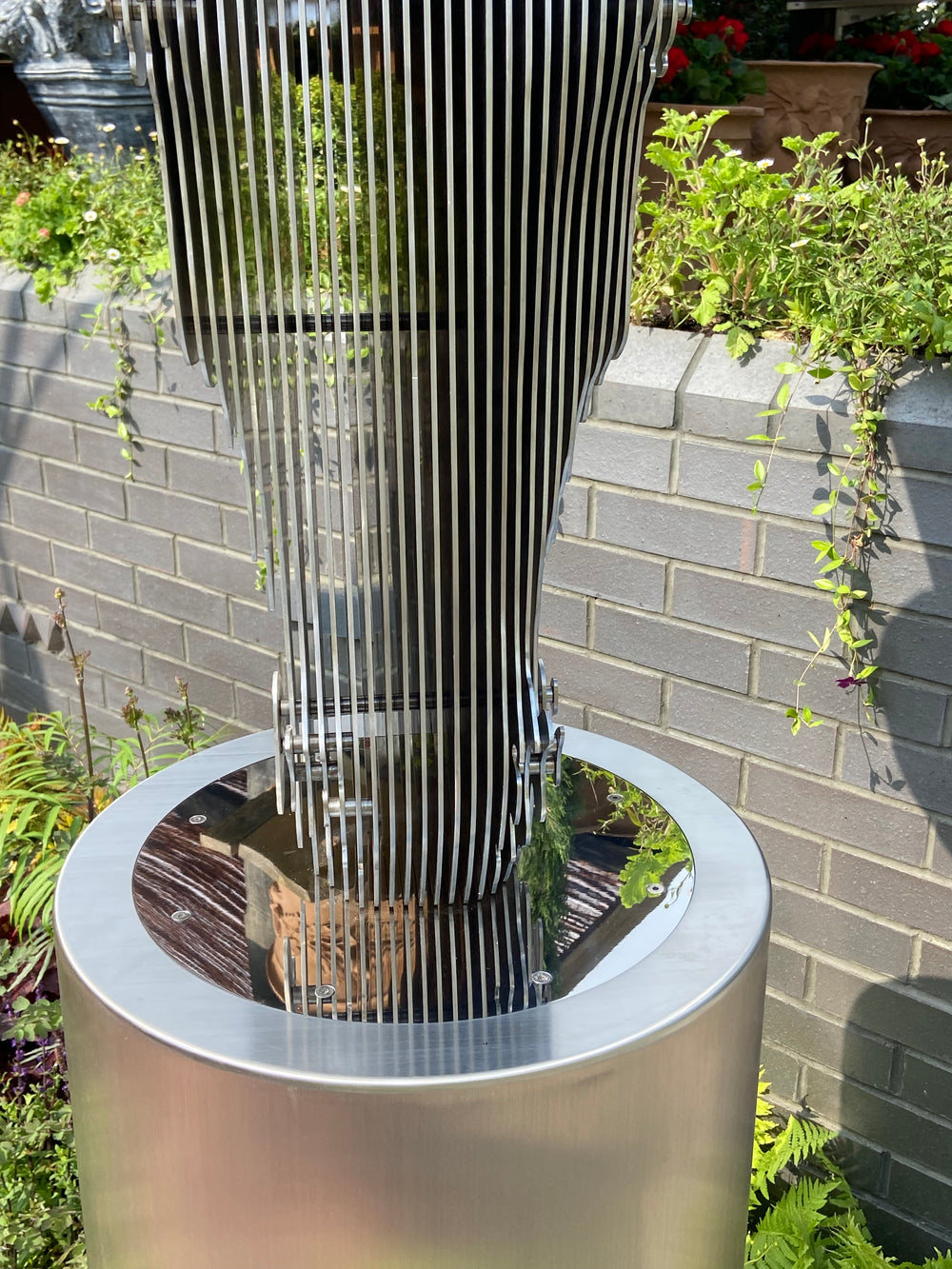 A sculptural stainless steel horse head by Tom Hiscox, featuring flowing contours and stylised form, displayed outdoors as a freestanding installation amid lush garden planting at the RHS Chelsea Flower Show, with rear views revealing the vertical steel slices and internal structure, culminating in a striking illuminated presentation set against the event’s floral and architectural backdrop.