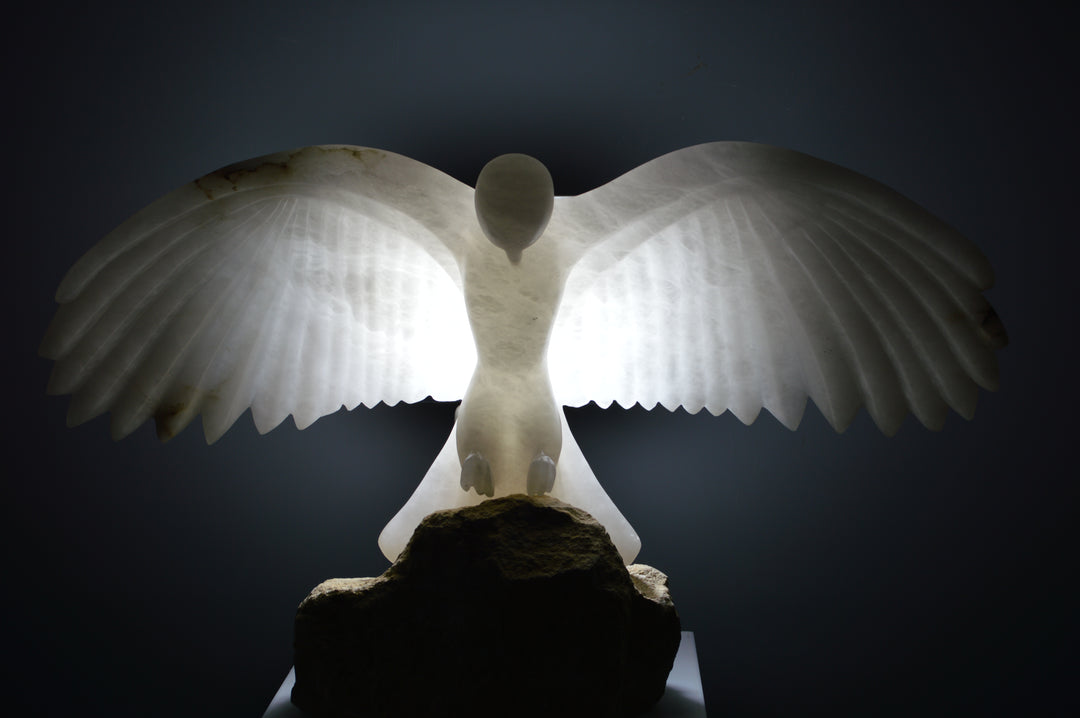 Deborah Harrison alabaster sculpture Caladrius, a translucent white bird with wide outstretched wings, finely carved feathers and smooth body, glowing with inner light on a rugged Farmington limestone base, symbolising purity, flight, and spiritual presence.