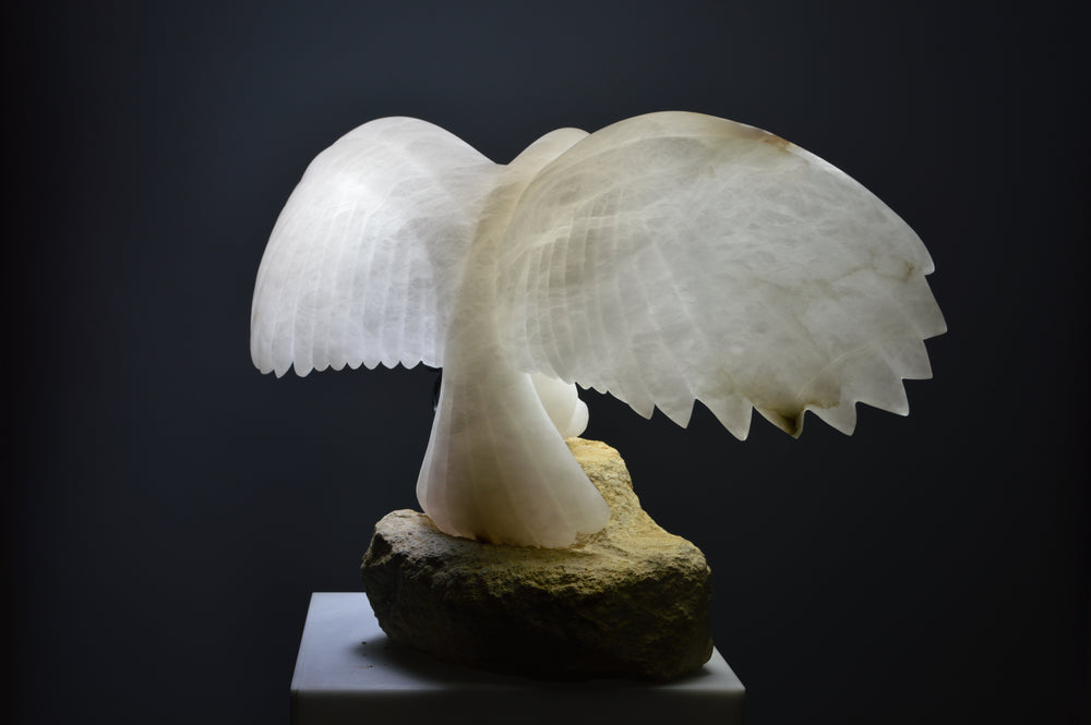 Deborah Harrison alabaster sculpture Caladrius, a translucent white bird with wide outstretched wings, finely carved feathers and smooth body, glowing with inner light on a rugged Farmington limestone base, symbolising purity, flight, and spiritual presence.