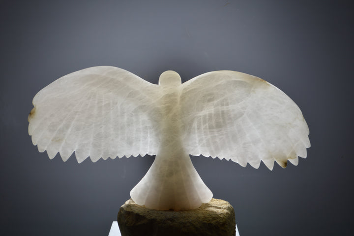 Deborah Harrison alabaster sculpture Caladrius, a translucent white bird with wide outstretched wings, finely carved feathers and smooth body, glowing with inner light on a rugged Farmington limestone base, symbolising purity, flight, and spiritual presence.