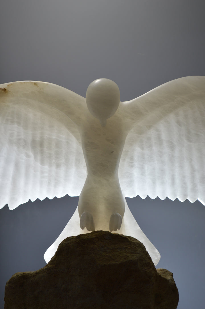 Deborah Harrison alabaster sculpture Caladrius, a translucent white bird with wide outstretched wings, finely carved feathers and smooth body, glowing with inner light on a rugged Farmington limestone base, symbolising purity, flight, and spiritual presence.
