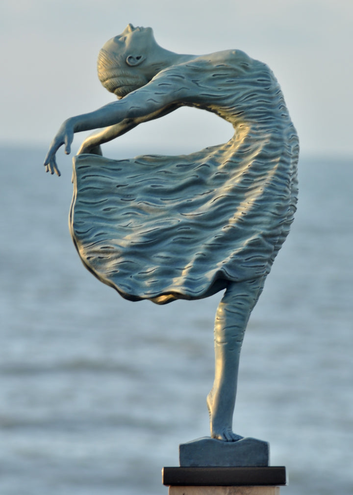 Bronze sculpture of a female figure leaning back in a flowing, wave-textured dress that echoes the motion of the sea, balanced delicately on one foot atop a plinth. Her arms and head are extended in a serene, open pose, with the sculpture photographed outdoors against a calm ocean horizon.
