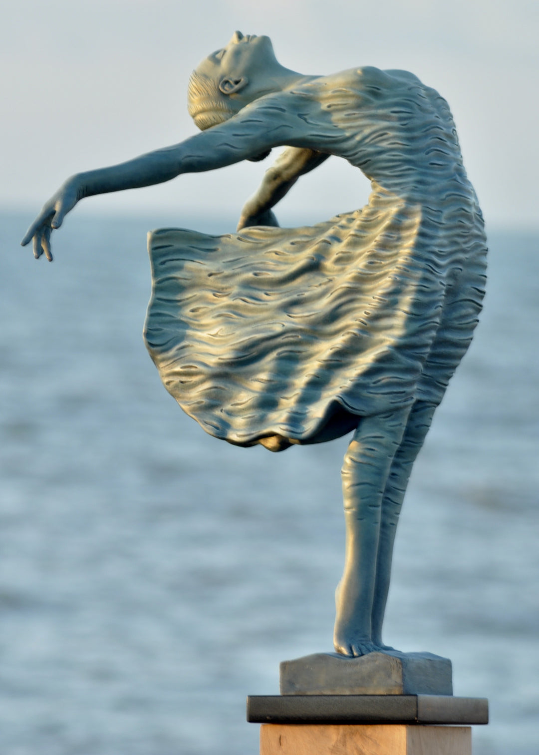 Bronze sculpture of a female figure leaning back in a flowing, wave-textured dress that echoes the motion of the sea, balanced delicately on one foot atop a plinth. Her arms and head are extended in a serene, open pose, with the sculpture photographed outdoors against a calm ocean horizon.