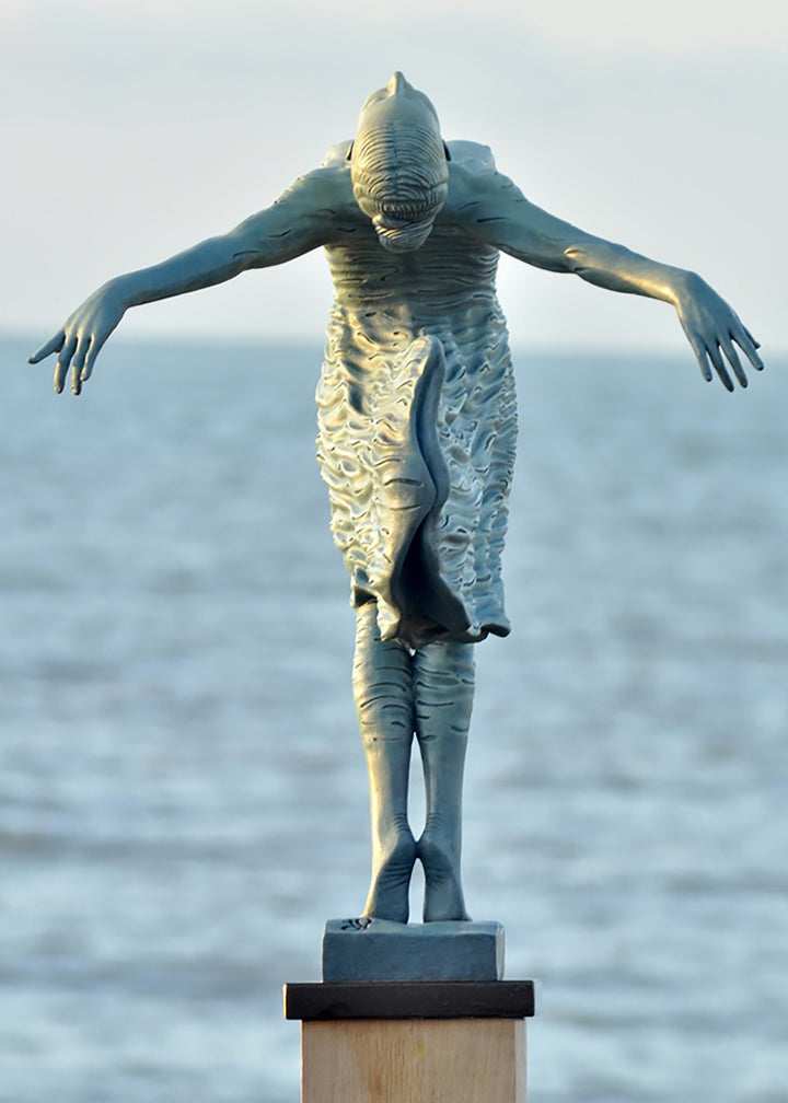 Bronze sculpture of a female figure leaning back in a flowing, wave-textured dress that echoes the motion of the sea, balanced delicately on one foot atop a plinth. Her arms and head are extended in a serene, open pose, with the sculpture photographed outdoors against a calm ocean horizon.