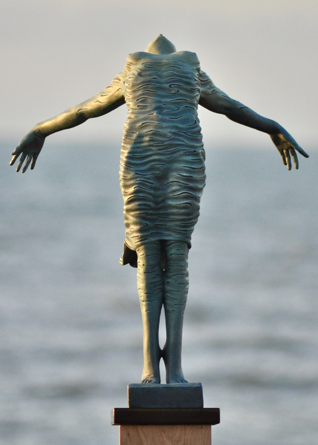 Bronze sculpture of a female figure leaning back in a flowing, wave-textured dress that echoes the motion of the sea, balanced delicately on one foot atop a plinth. Her arms and head are extended in a serene, open pose, with the sculpture photographed outdoors against a calm ocean horizon.