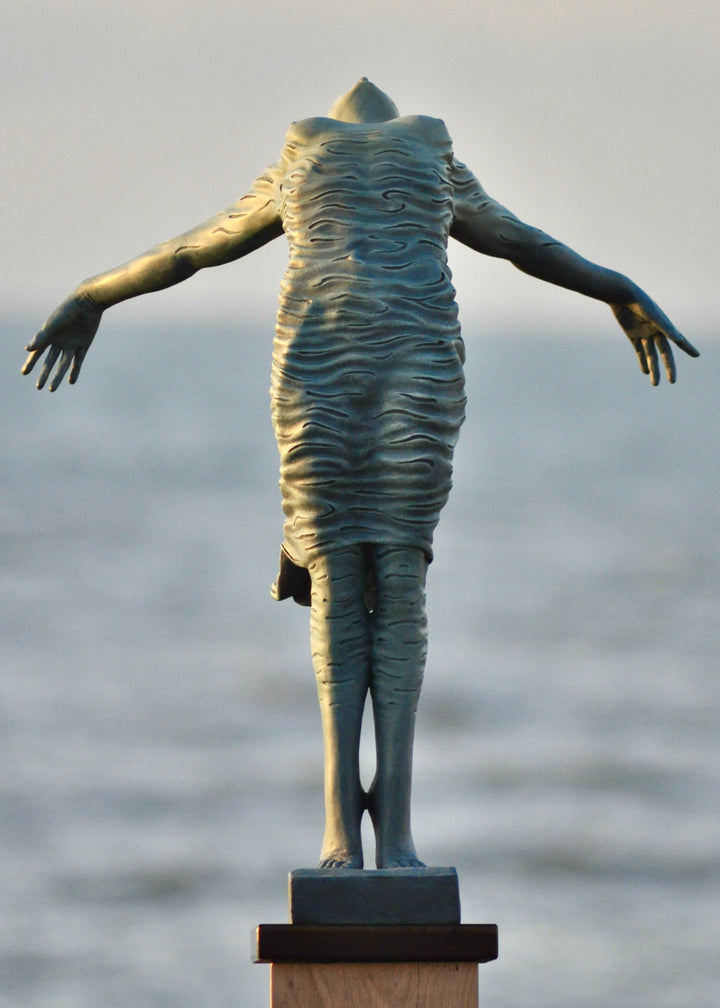 Bronze sculpture of a female figure leaning back in a flowing, wave-textured dress that echoes the motion of the sea, balanced delicately on one foot atop a plinth. Her arms and head are extended in a serene, open pose, with the sculpture photographed outdoors against a calm ocean horizon.