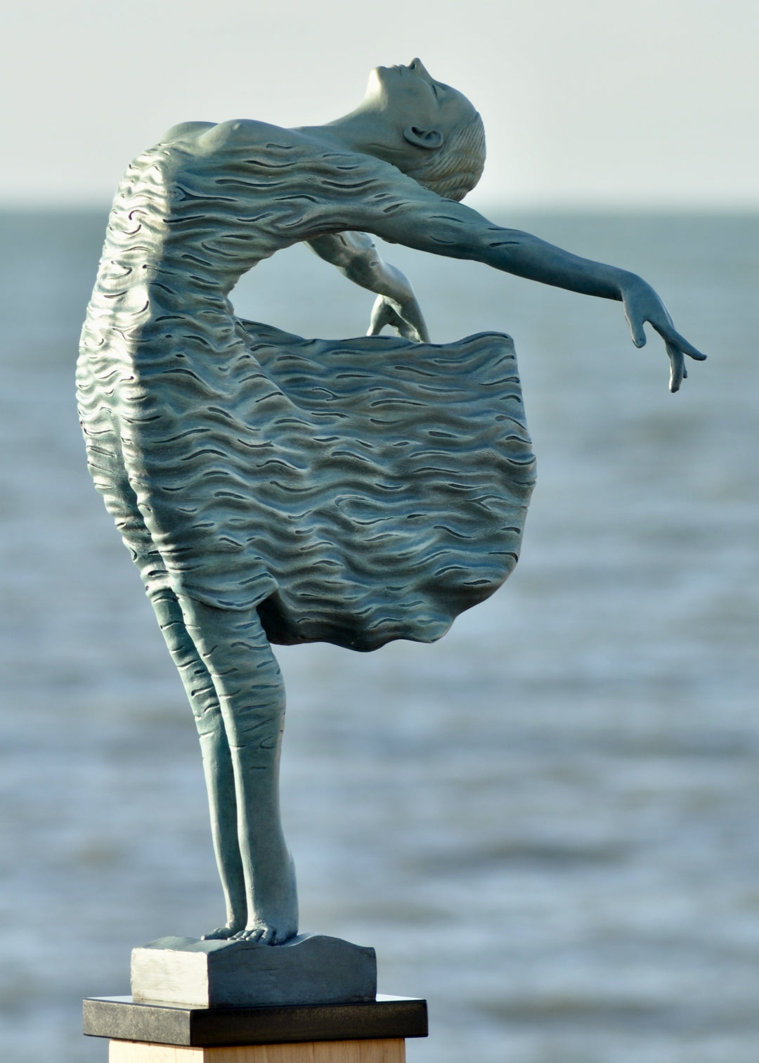Bronze sculpture of a female figure leaning back in a flowing, wave-textured dress that echoes the motion of the sea, balanced delicately on one foot atop a plinth. Her arms and head are extended in a serene, open pose, with the sculpture photographed outdoors against a calm ocean horizon.