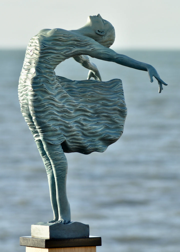 Bronze sculpture of a female figure leaning back in a flowing, wave-textured dress that echoes the motion of the sea, balanced delicately on one foot atop a plinth. Her arms and head are extended in a serene, open pose, with the sculpture photographed outdoors against a calm ocean horizon.