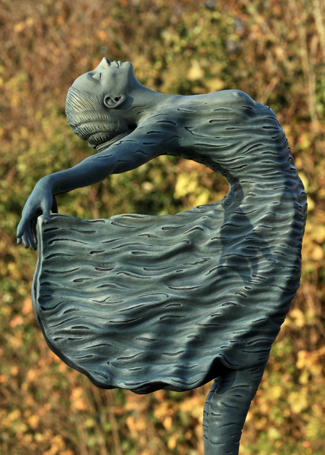 Bronze sculpture of a female figure leaning back in a flowing, wave-textured dress that echoes the motion of the sea, balanced delicately on one foot atop a plinth. Her arms and head are extended in a serene, open pose, with the sculpture photographed outdoors against a calm ocean horizon.