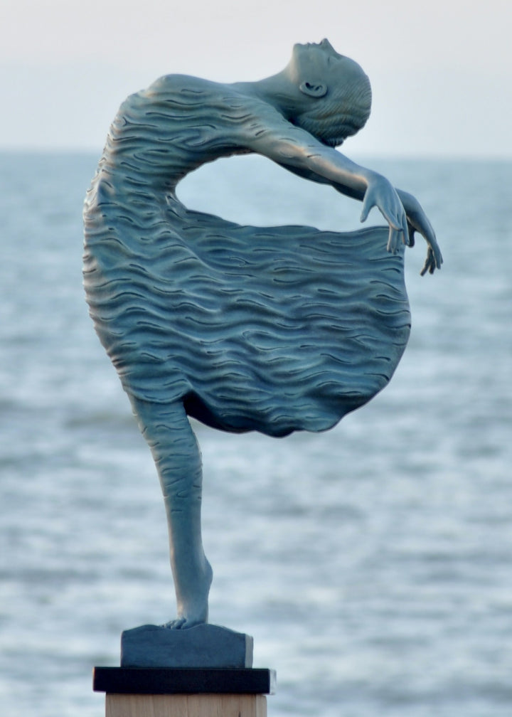 Bronze sculpture of a female figure leaning back in a flowing, wave-textured dress that echoes the motion of the sea, balanced delicately on one foot atop a plinth. Her arms and head are extended in a serene, open pose, with the sculpture photographed outdoors against a calm ocean horizon.