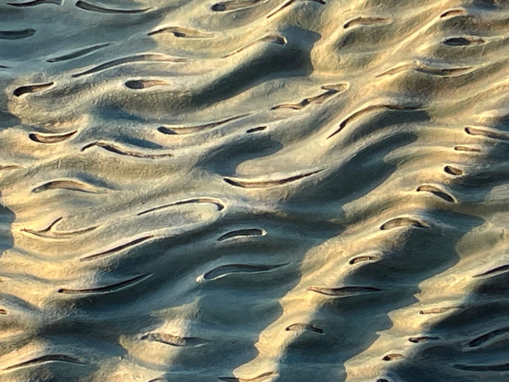 Bronze sculpture of a female figure leaning back in a flowing, wave-textured dress that echoes the motion of the sea, balanced delicately on one foot atop a plinth. Her arms and head are extended in a serene, open pose, with the sculpture photographed outdoors against a calm ocean horizon.