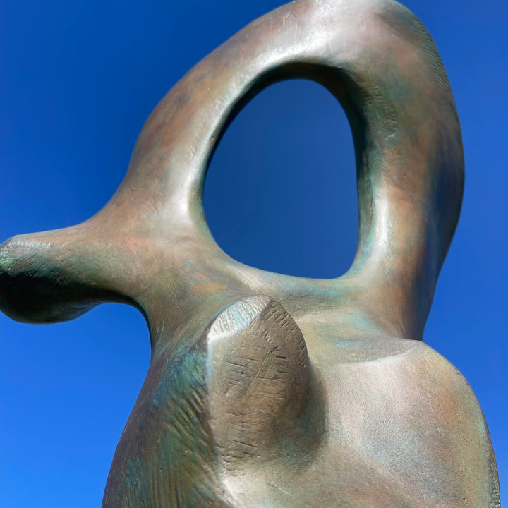 Abstract bronze sculpture of a stylised female torso with flowing curves and a hollowed circular form near the head, displayed in various outdoor and indoor settings. The sculpture has a rich patinated surface with green, gold, and bronze tones.