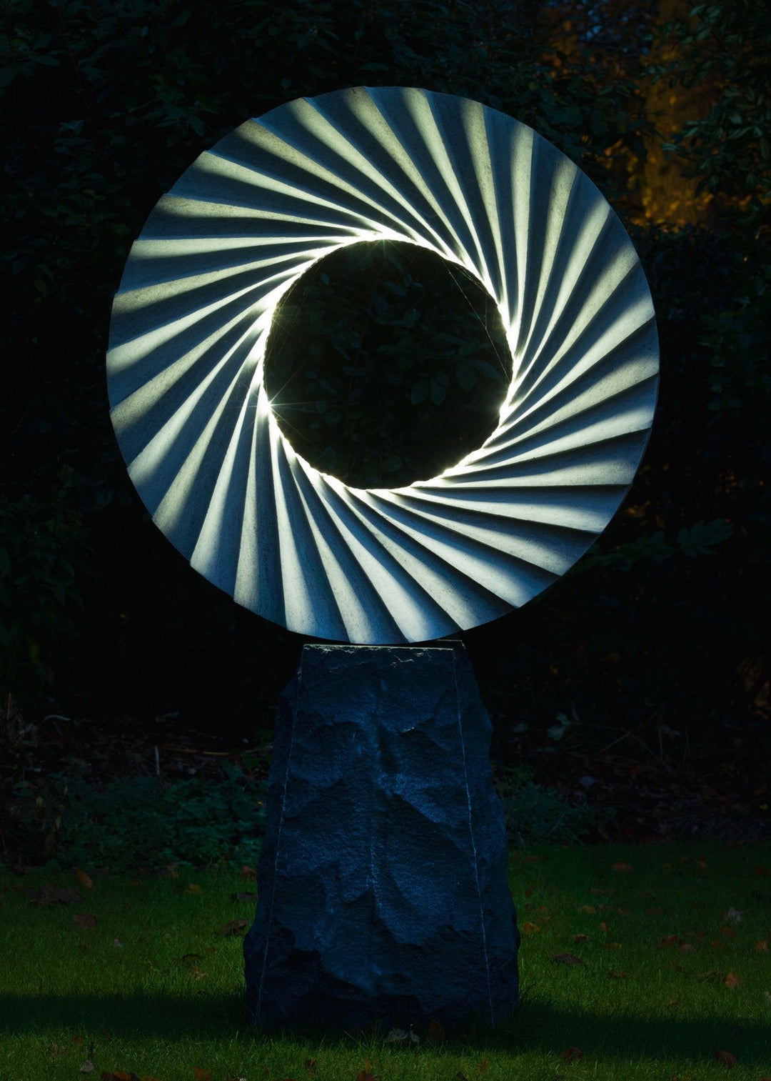 ‘Catherine Wheel VIII’ by Nicolas Moreton MRSS, a unique contemporary abstract stone sculpture carved from Ancaster Weatherbed Limestone, featuring a large circular form with a central opening surrounded by precisely carved, twisting radial ridges that create a spiralling pattern. The pale grey and cream limestone contrasts with the rough, dark charcoal stone pedestal. Integrated LED lighting illuminates the inner edges of the spiral with bright white light, creating alternating bands of light and shadow. P