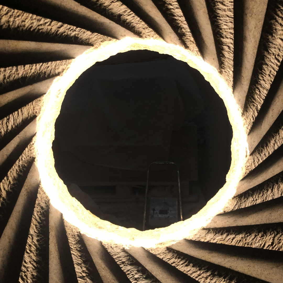 ‘Catherine Wheel VIII’ by Nicolas Moreton MRSS, a unique contemporary abstract stone sculpture carved from Ancaster Weatherbed Limestone, featuring a large circular form with a central opening surrounded by precisely carved, twisting radial ridges that create a spiralling pattern. The pale grey and cream limestone contrasts with the rough, dark charcoal stone pedestal. Integrated LED lighting illuminates the inner edges of the spiral with bright white light, creating alternating bands of light and shadow. P