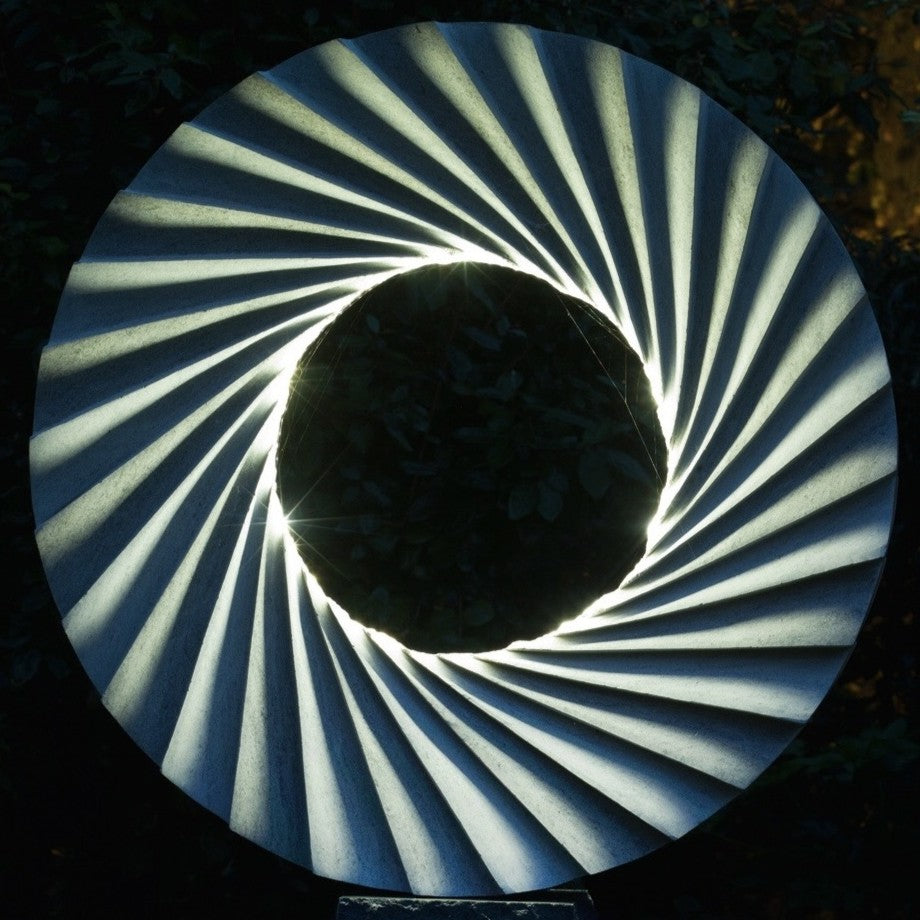 ‘Catherine Wheel VIII’ by Nicolas Moreton MRSS, a unique contemporary abstract stone sculpture carved from Ancaster Weatherbed Limestone, featuring a large circular form with a central opening surrounded by precisely carved, twisting radial ridges that create a spiralling pattern. The pale grey and cream limestone contrasts with the rough, dark charcoal stone pedestal. Integrated LED lighting illuminates the inner edges of the spiral with bright white light, creating alternating bands of light and shadow. P