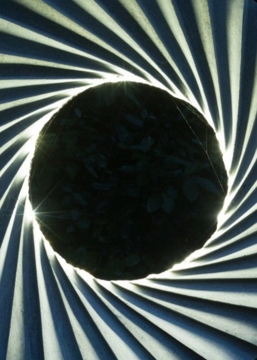 ‘Catherine Wheel VIII’ by Nicolas Moreton MRSS, a unique contemporary abstract stone sculpture carved from Ancaster Weatherbed Limestone, featuring a large circular form with a central opening surrounded by precisely carved, twisting radial ridges that create a spiralling pattern. The pale grey and cream limestone contrasts with the rough, dark charcoal stone pedestal. Integrated LED lighting illuminates the inner edges of the spiral with bright white light, creating alternating bands of light and shadow. P