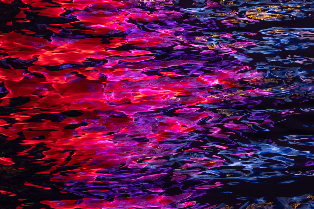 Vibrant abstract photograph capturing multicoloured neon reflections on water, with bold swirls of red, pink, purple, blue, and gold set against a dark background.