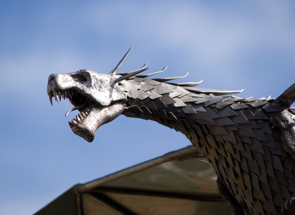 An impressive kinetic dragon sculpture by Martin Jarvis, made entirely from polished and textured metal. The dragon is mounted on a sweeping, curved arm that allows it to pivot and move with the wind. The body is detailed with overlapping metal scales, while the expansive wings—crafted from dark, matte-finished panels—give the illusion of flight. The sculpture captures dynamic energy and motion, designed to gently sway or turn with air currents. Positioned outdoors against blue skies, the piece