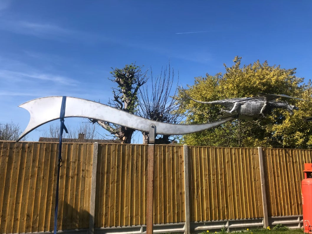 An impressive kinetic dragon sculpture by Martin Jarvis, made entirely from polished and textured metal. The dragon is mounted on a sweeping, curved arm that allows it to pivot and move with the wind. The body is detailed with overlapping metal scales, while the expansive wings—crafted from dark, matte-finished panels—give the illusion of flight. The sculpture captures dynamic energy and motion, designed to gently sway or turn with air currents. Positioned outdoors against blue skies, the piece