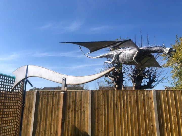 An impressive kinetic dragon sculpture by Martin Jarvis, made entirely from polished and textured metal. The dragon is mounted on a sweeping, curved arm that allows it to pivot and move with the wind. The body is detailed with overlapping metal scales, while the expansive wings—crafted from dark, matte-finished panels—give the illusion of flight. The sculpture captures dynamic energy and motion, designed to gently sway or turn with air currents. Positioned outdoors against blue skies, the piece