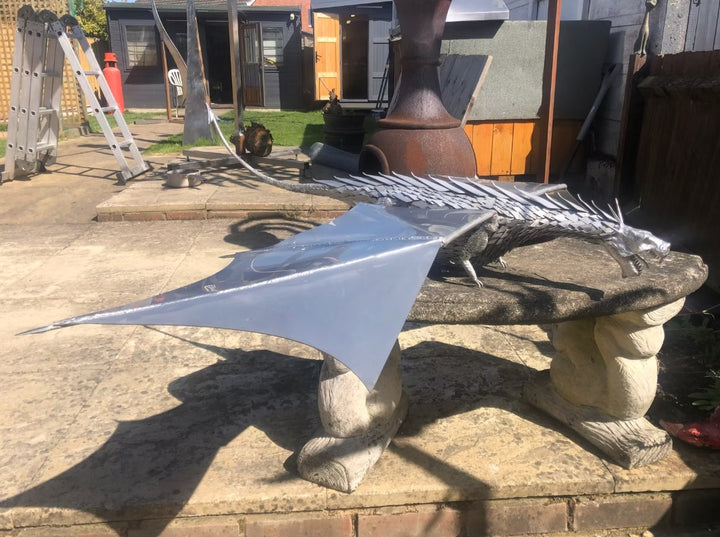 An impressive kinetic dragon sculpture by Martin Jarvis, made entirely from polished and textured metal. The dragon is mounted on a sweeping, curved arm that allows it to pivot and move with the wind. The body is detailed with overlapping metal scales, while the expansive wings—crafted from dark, matte-finished panels—give the illusion of flight. The sculpture captures dynamic energy and motion, designed to gently sway or turn with air currents. Positioned outdoors against blue skies, the piece