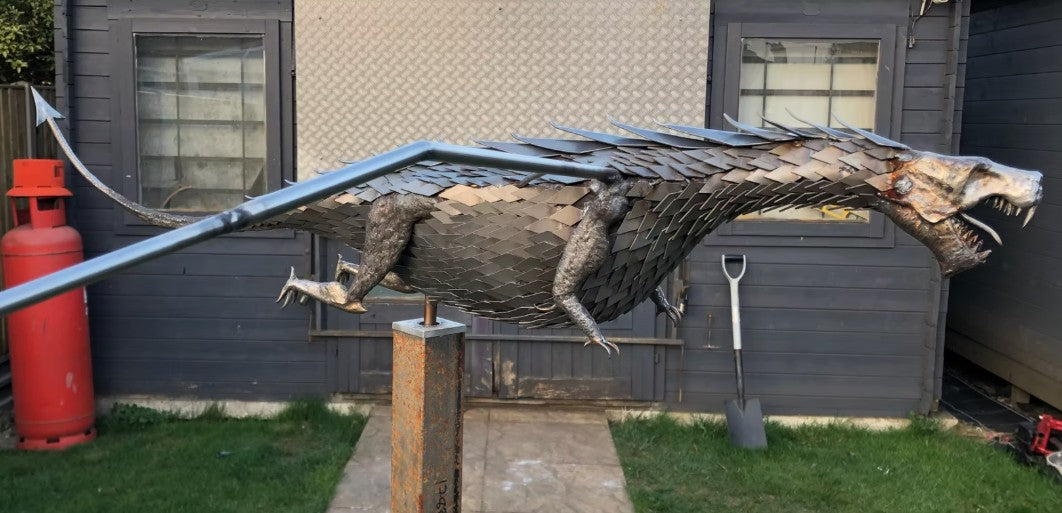 An impressive kinetic dragon sculpture by Martin Jarvis, made entirely from polished and textured metal. The dragon is mounted on a sweeping, curved arm that allows it to pivot and move with the wind. The body is detailed with overlapping metal scales, while the expansive wings—crafted from dark, matte-finished panels—give the illusion of flight. The sculpture captures dynamic energy and motion, designed to gently sway or turn with air currents. Positioned outdoors against blue skies, the piece
