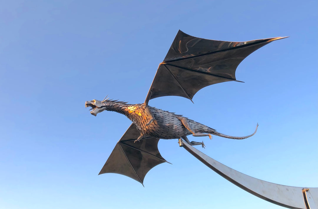 An impressive kinetic dragon sculpture by Martin Jarvis, made entirely from polished and textured metal. The dragon is mounted on a sweeping, curved arm that allows it to pivot and move with the wind. The body is detailed with overlapping metal scales, while the expansive wings—crafted from dark, matte-finished panels—give the illusion of flight. The sculpture captures dynamic energy and motion, designed to gently sway or turn with air currents. Positioned outdoors against blue skies, the piece