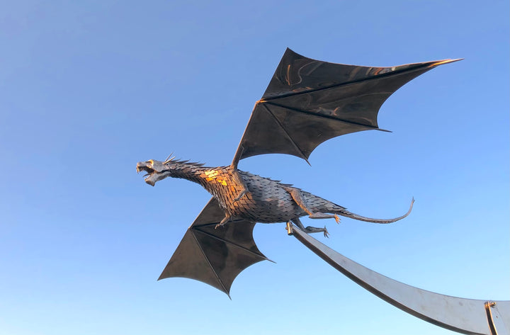 An impressive kinetic dragon sculpture by Martin Jarvis, made entirely from polished and textured metal. The dragon is mounted on a sweeping, curved arm that allows it to pivot and move with the wind. The body is detailed with overlapping metal scales, while the expansive wings—crafted from dark, matte-finished panels—give the illusion of flight. The sculpture captures dynamic energy and motion, designed to gently sway or turn with air currents. Positioned outdoors against blue skies, the piece