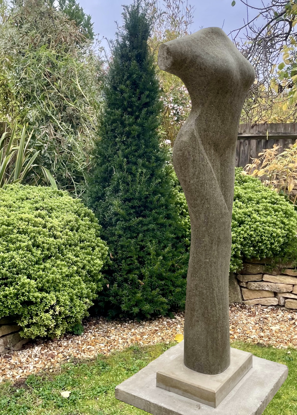 Tall stone sculpture by Martin Cash titled Chasing the Curve, hand-carved in Ancaster Blue Stone with a Purbeck Stone base. The piece depicts an abstract female torso with graceful curves and smooth contours, designed as a contemporary outdoor sculpture for gardens or public spaces.