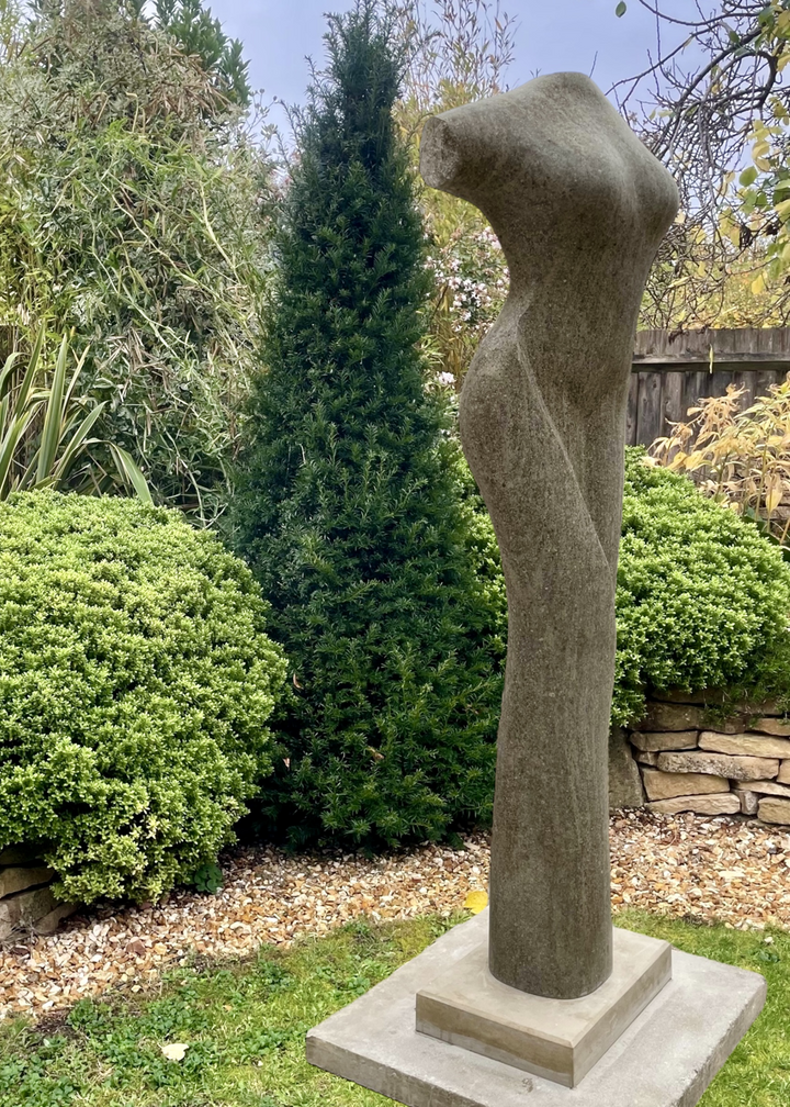 Tall stone sculpture by Martin Cash titled Chasing the Curve, hand-carved in Ancaster Blue Stone with a Purbeck Stone base. The piece depicts an abstract female torso with graceful curves and smooth contours, designed as a contemporary outdoor sculpture for gardens or public spaces.