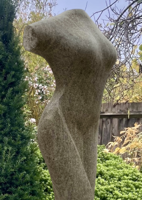 Tall stone sculpture by Martin Cash titled Chasing the Curve, hand-carved in Ancaster Blue Stone with a Purbeck Stone base. The piece depicts an abstract female torso with graceful curves and smooth contours, designed as a contemporary outdoor sculpture for gardens or public spaces.