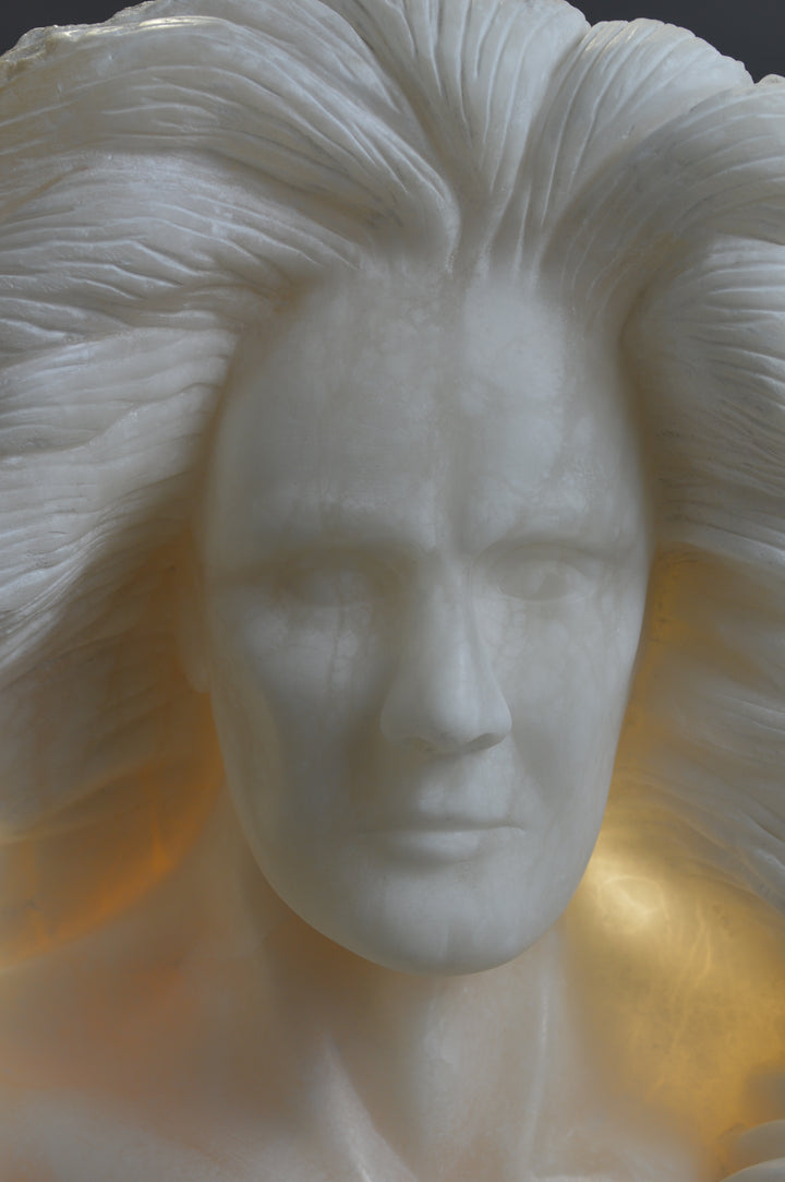 Deborah Harrison alabaster sculpture Cherubim, one-off hand-carved work in glowing white alabaster with translucent qualities, depicting a powerful male figure surrounded by symbolic forms including a goat-like animal, a bird, and a lion, mounted on slate, blending myth, spirituality, and masterful stone carving.