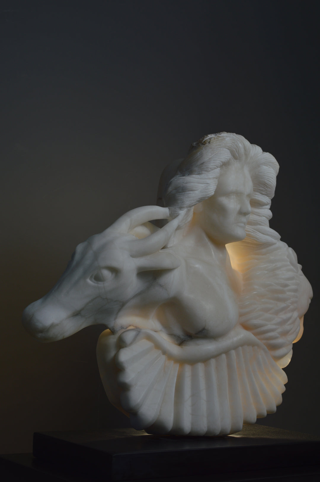 Deborah Harrison alabaster sculpture Cherubim, one-off hand-carved work in glowing white alabaster with translucent qualities, depicting a powerful male figure surrounded by symbolic forms including a goat-like animal, a bird, and a lion, mounted on slate, blending myth, spirituality, and masterful stone carving.