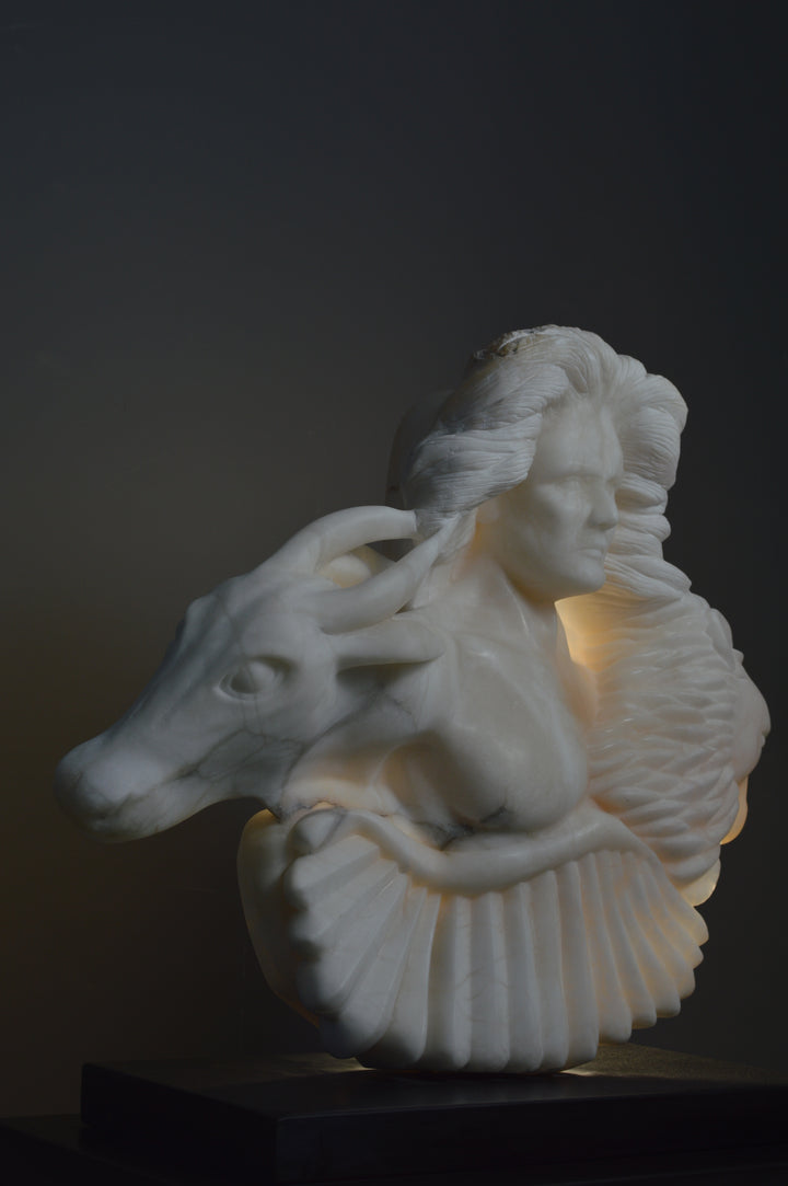 Deborah Harrison alabaster sculpture Cherubim, one-off hand-carved work in glowing white alabaster with translucent qualities, depicting a powerful male figure surrounded by symbolic forms including a goat-like animal, a bird, and a lion, mounted on slate, blending myth, spirituality, and masterful stone carving.