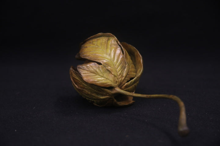 Bronze sculpture of a curled-up dormouse nestled inside a detailed horse chestnut leaf, with a long stem extending outward, set against a dark background.