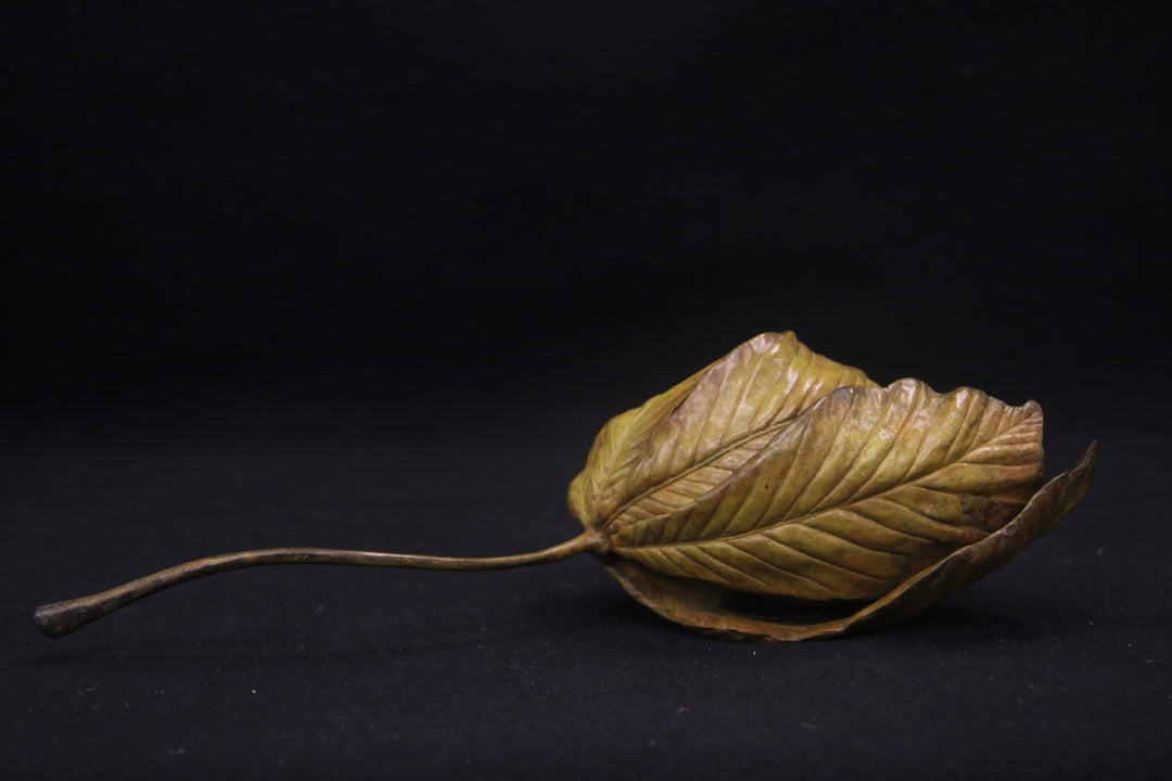 Bronze sculpture of a curled-up dormouse nestled inside a detailed horse chestnut leaf, with a long stem extending outward, set against a dark background.