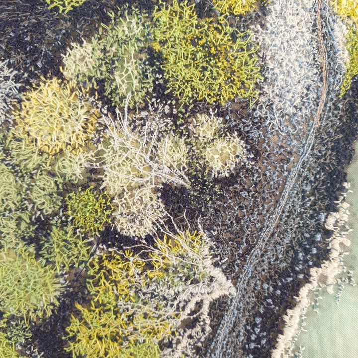 Aerial-style embroidered artwork of a rugged coastline with turquoise water, rocky textures, and mossy vegetation in shades of green and grey.

