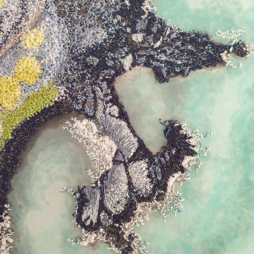 Aerial-style embroidered artwork of a rugged coastline with turquoise water, rocky textures, and mossy vegetation in shades of green and grey.
