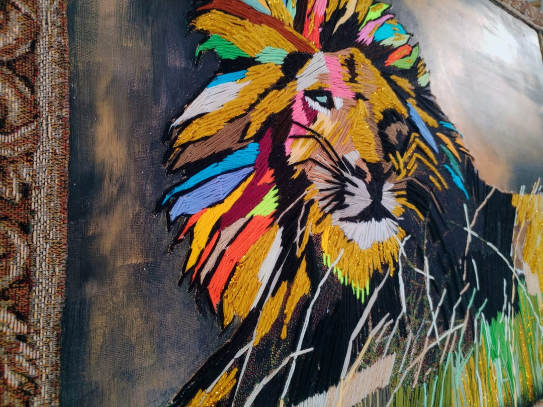 “Comeback” by Helena Safari, hand embroidery on canvas. A majestic lion reclines in tall grass, its mane bursting with embroidered colour – fiery yellows, radiant oranges, electric blues, vivid pinks, and deep greens against a golden-yellow and teal sky. The animal’s intense gaze and flowing mane exude both strength and renewal, while the textured stitches give the work depth and vitality.
