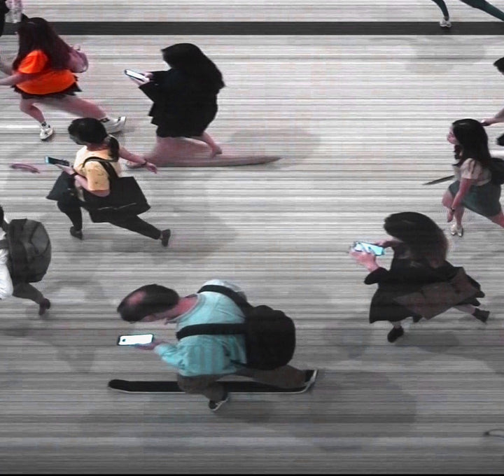 High-angle composite image of Hong Kong commuters moving through a brightly lit station, captured using high-speed video reconstruction. The figures are partially blurred and distorted, their faces downturned toward phones—creating a visual rhythm that blurs individuality, echoing the dehumanising pace of mass transit systems.